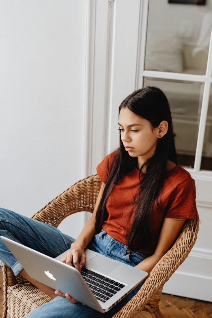 pexels-photo-7281588-7281588 Young girl comfortably sitting indoors, focused on her laptop for casual browsing or online activity.
