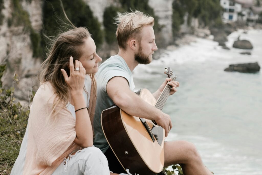 Side view of young bearded male musician playing guitar while sitting on high cliff with girlfriend and enjoying ocean view