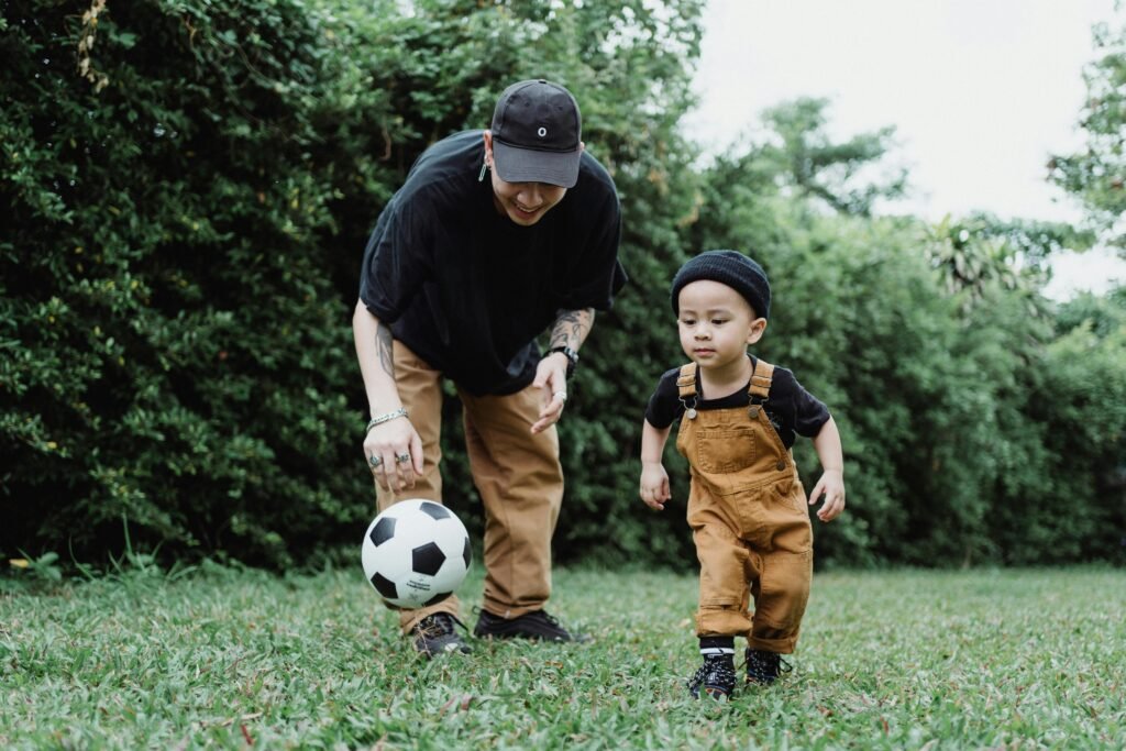 pexels-photo-4933837-4933837 Father and young son enjoying playing soccer together in a lush green park.