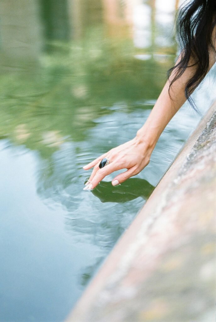 A close-up of a woman's hand with a ring gently touching the calm water surface.
